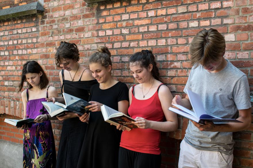 vijf jongeren lezen een boek, ze staan naast elkaar en leunen tegen een bakstenen muur